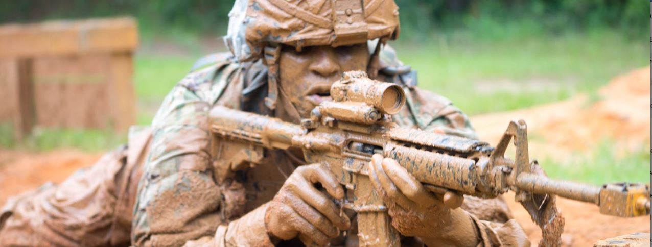 soldier covered in mud aiming a rifle in the prone position